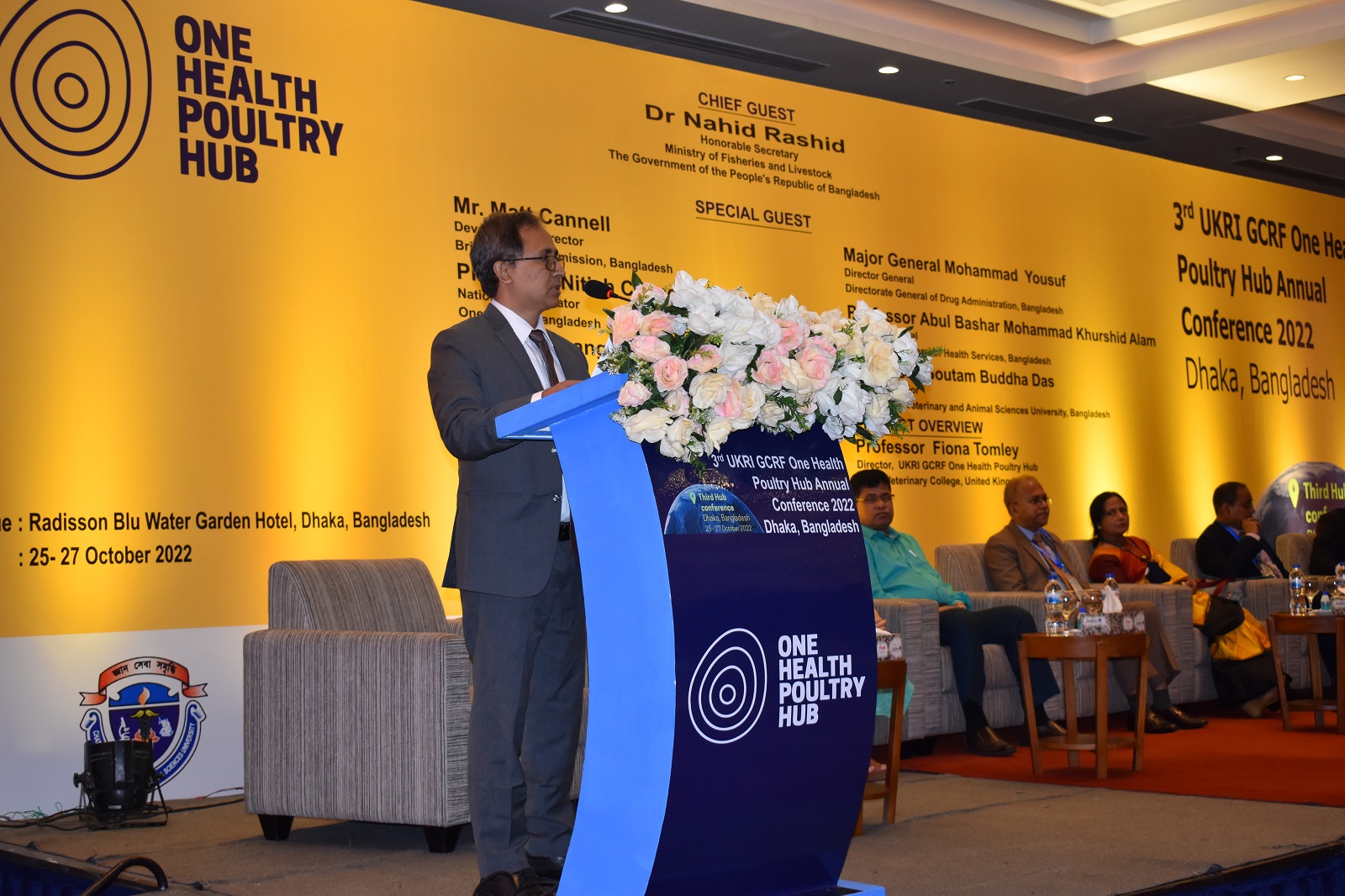 Prof Hoque at a podium beihnd a large Hub nanner detailing the conference speakers.