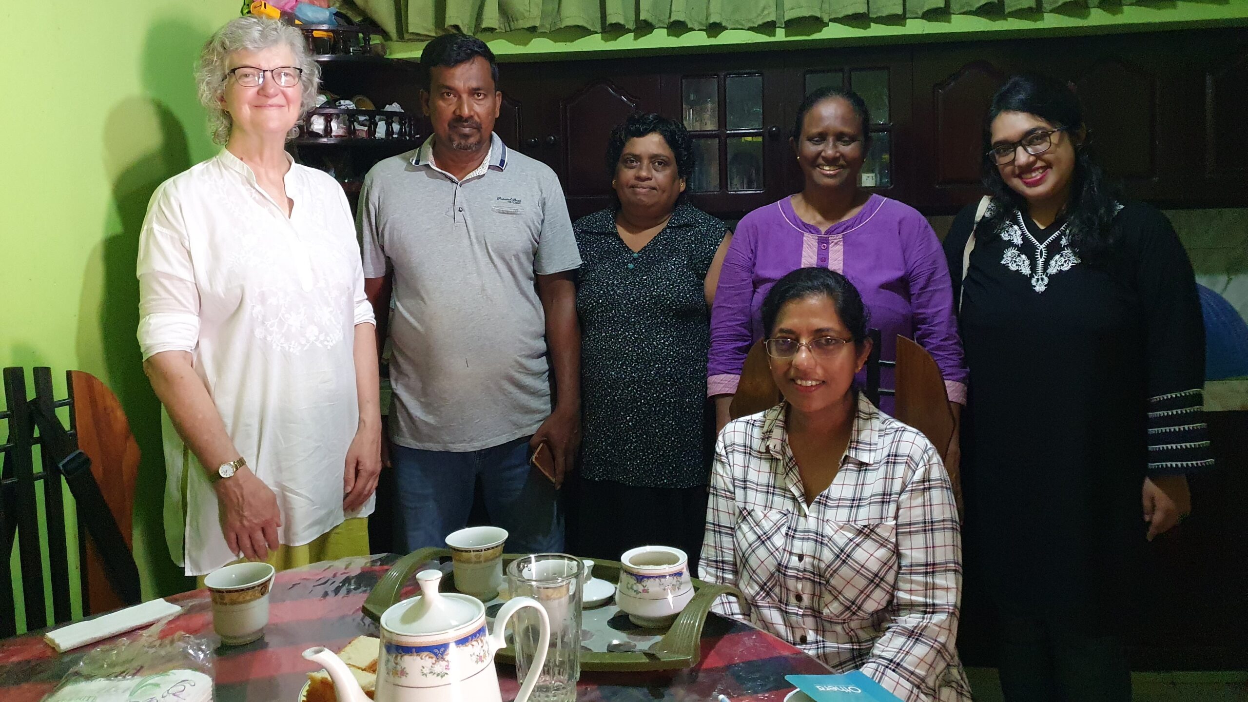 Six people, five standing, around a table lade with cups and a teapot