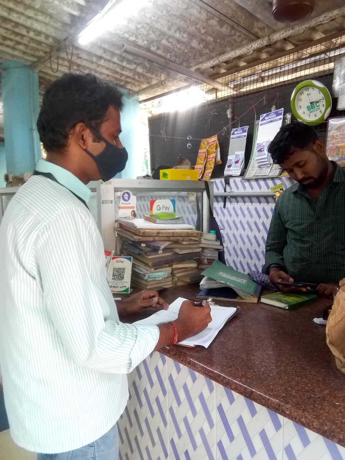 man at shop counter in mask, with another man looking at a mobile phone